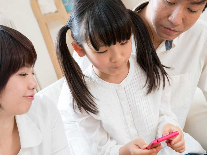 小学生がスマホをいじるのを見ているお母さんとお父さん