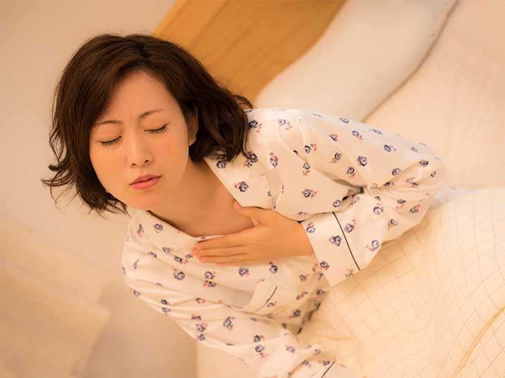 動悸やだるさで眠れず胸をさすりながらベッドに座っている女性